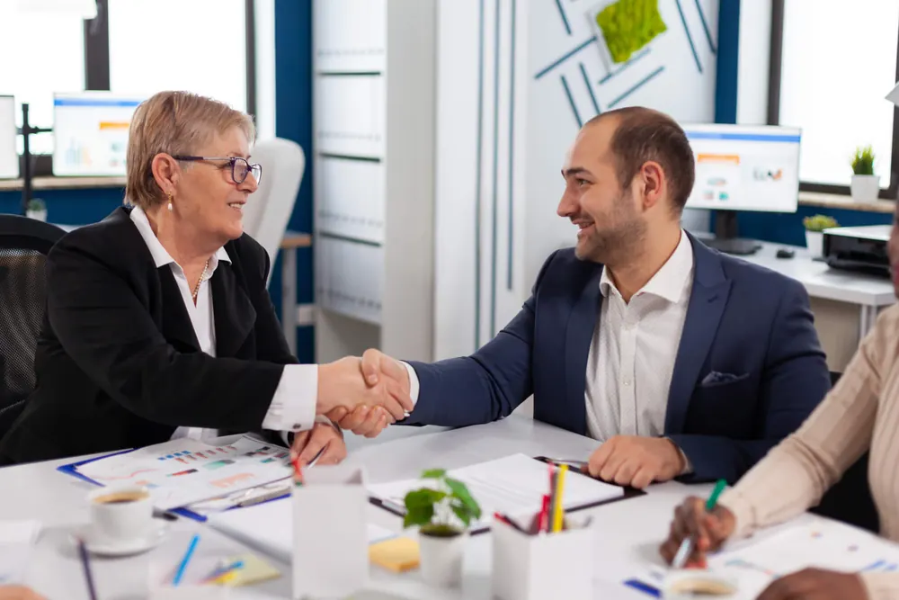 Two people shaking hands across a desk at a business meeting